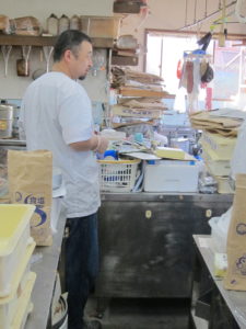 Toshi-san is in his factory, smiling. He's facing towards the sink, getting ready to wash some mochi-making equipment.