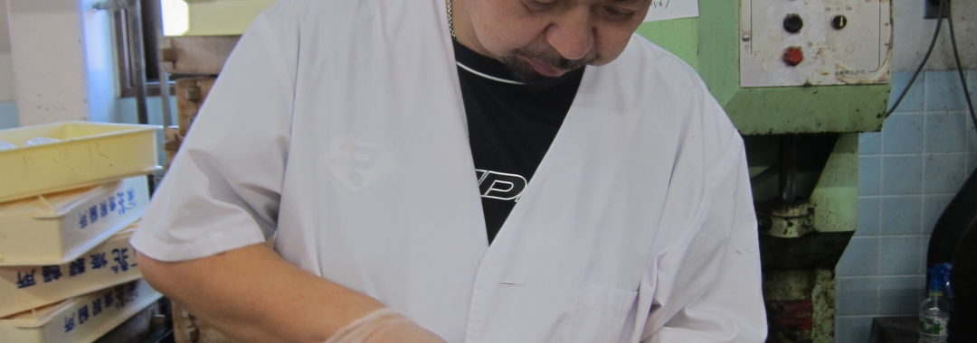 Toshi-san cuts into a daifuku mochi with a knife. He is standing in his factory.