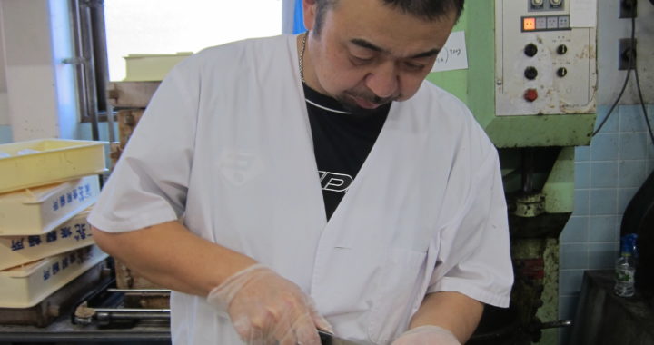 Toshi-san cuts into a daifuku mochi with a knife. He is standing in his factory.