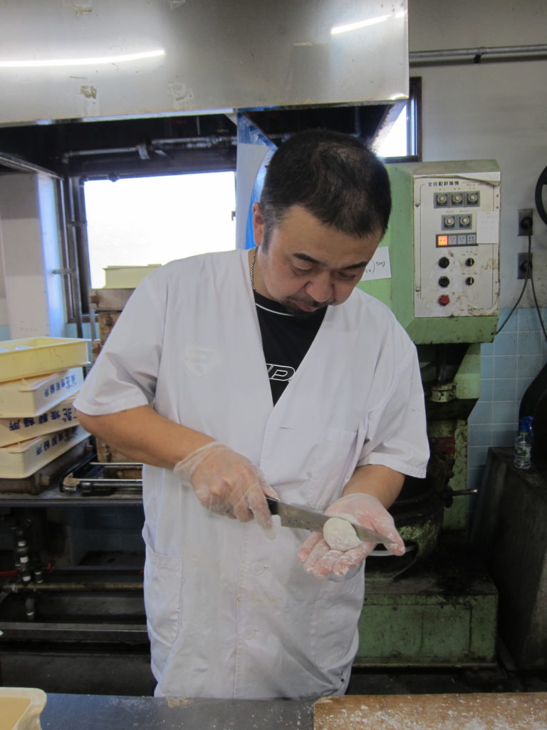 Toshi-san cuts into a daifuku mochi with a knife. He is standing in his factory.