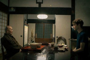 On the left, Shouhouji's chief abbot, and Alex on the right are deep in conversation. They sit across from each other at a dark wood table with light handmade teaware on it.
