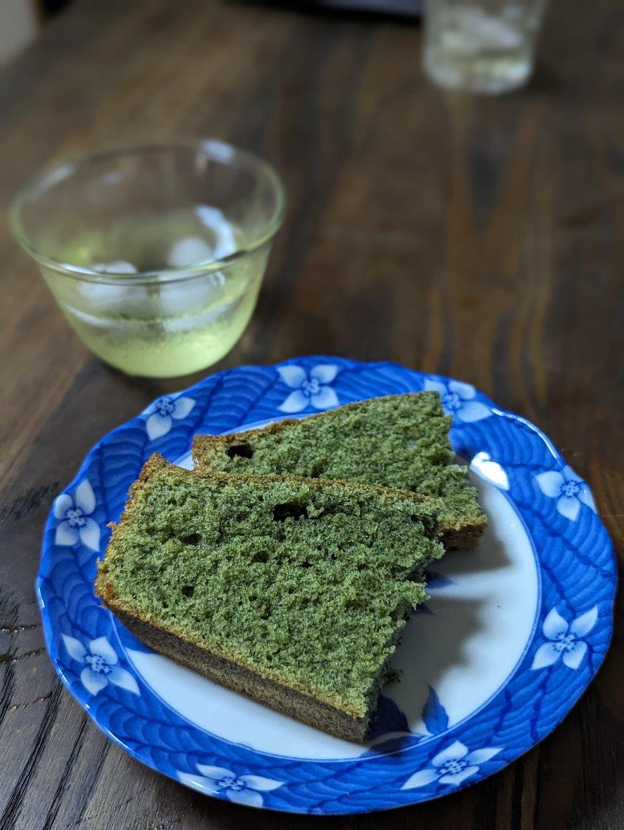 2 slices of green mugwort cake without crumble on a white and blue plate, with a glass of green tea in the background